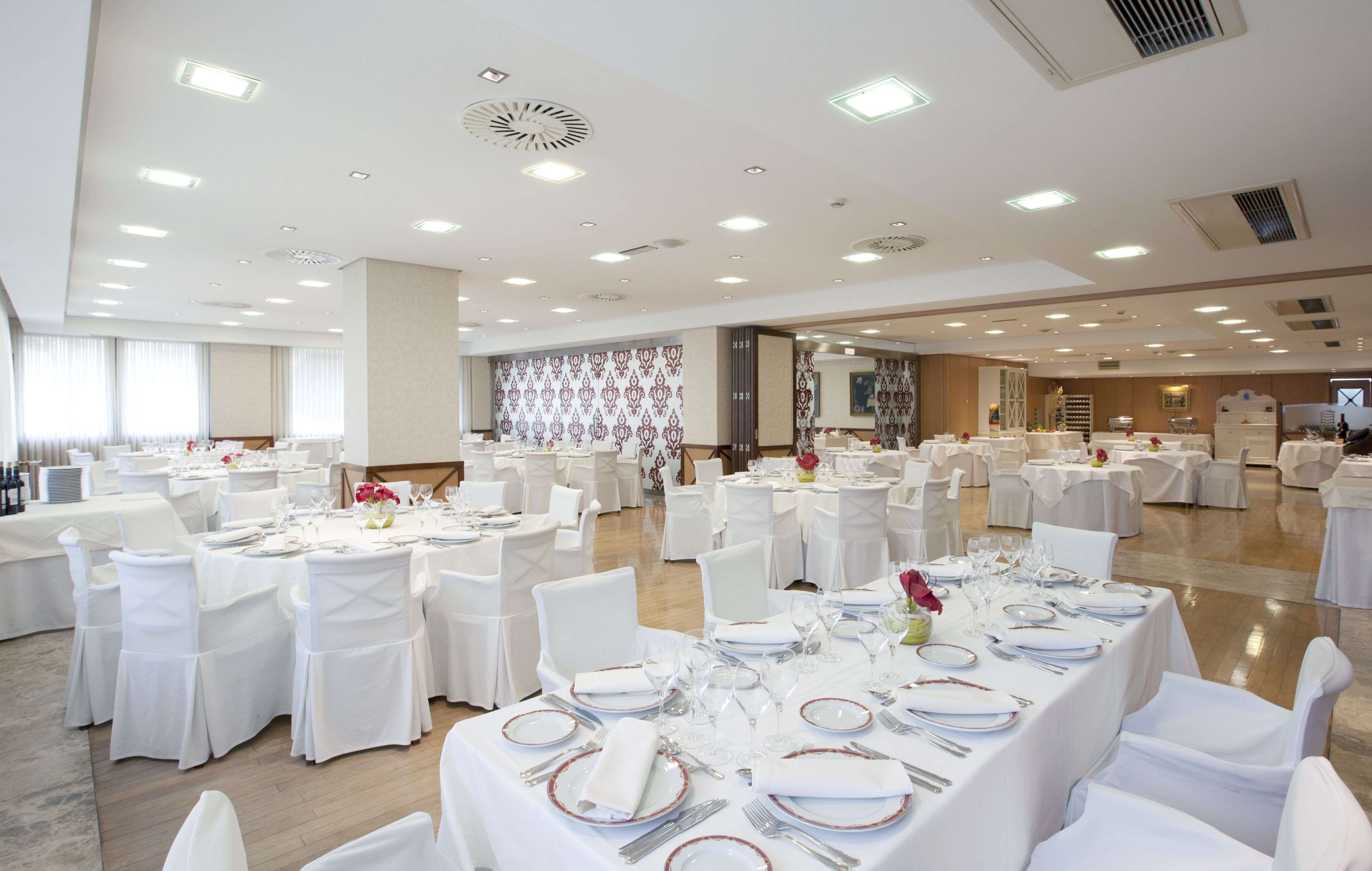 Un amplio salón de banquetes con varias mesas redondas dispuestas para comer, sillas blancas e iluminación en el techo.