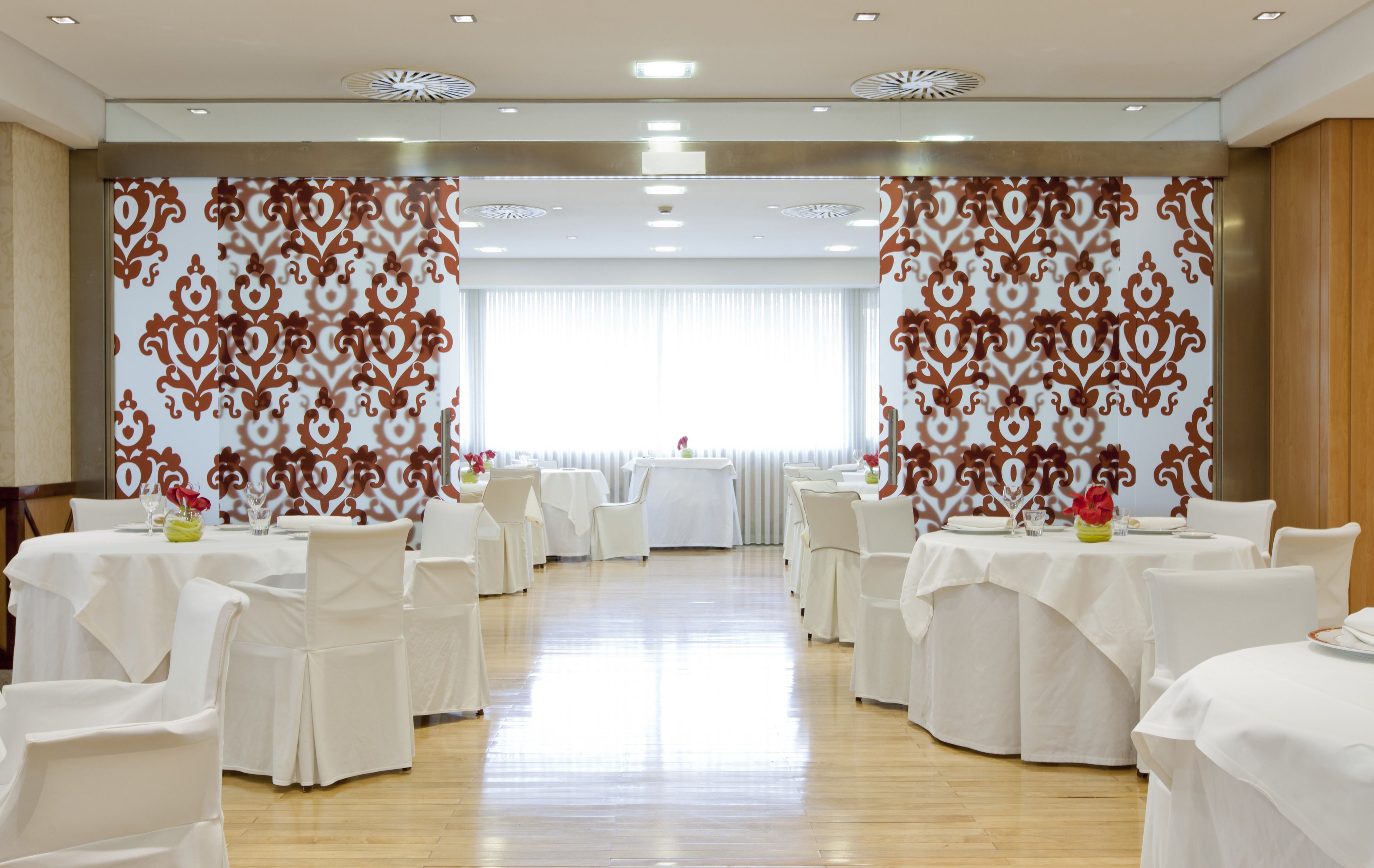 A banquet hall with round tables and white chairs, a wooden floor, and patterned curtains.
