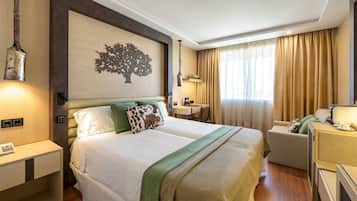 Una habitación de hotel con cama, mesitas de noche, escritorio, una silla y un ventanal con cortinas.