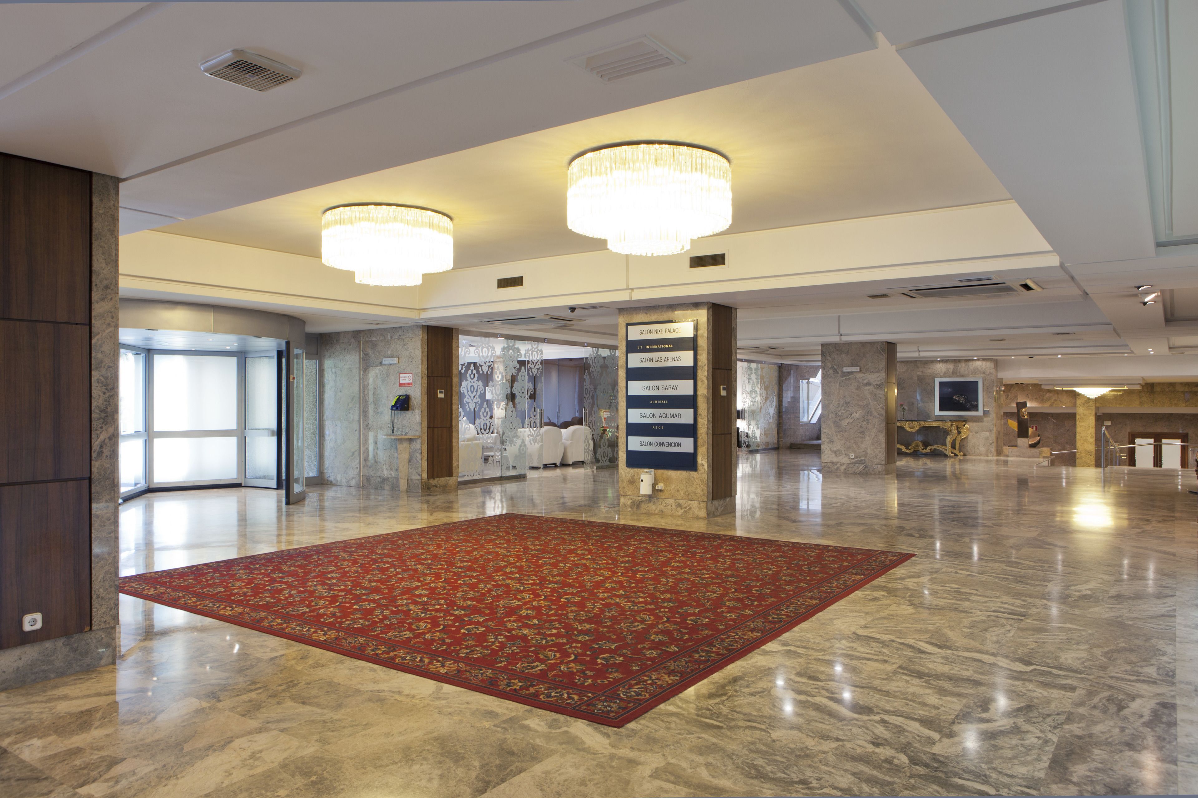 Um hall amplo com carpete vermelho, lustres, piso de mármore e um sinal que indica a localização de diversos serviços do hotel.