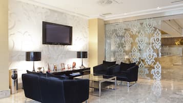 Eine moderne Lobby mit einem an der Wand befestigten Flachbildfernseher, einem Glastisch und zwei schwarzen Sofas, sowie einem dekorativen Glaseinsatz.