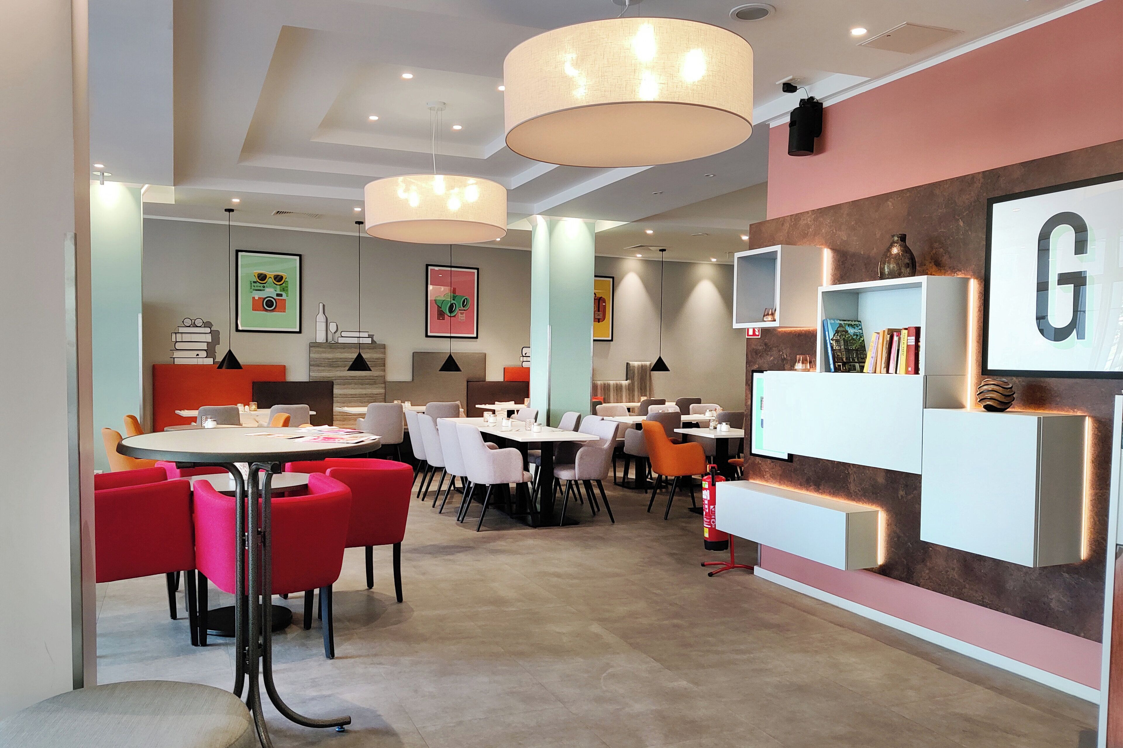 A modern dining area with round tables and chairs, pendant lighting, and a wall-mounted shelf with books and decorative items.