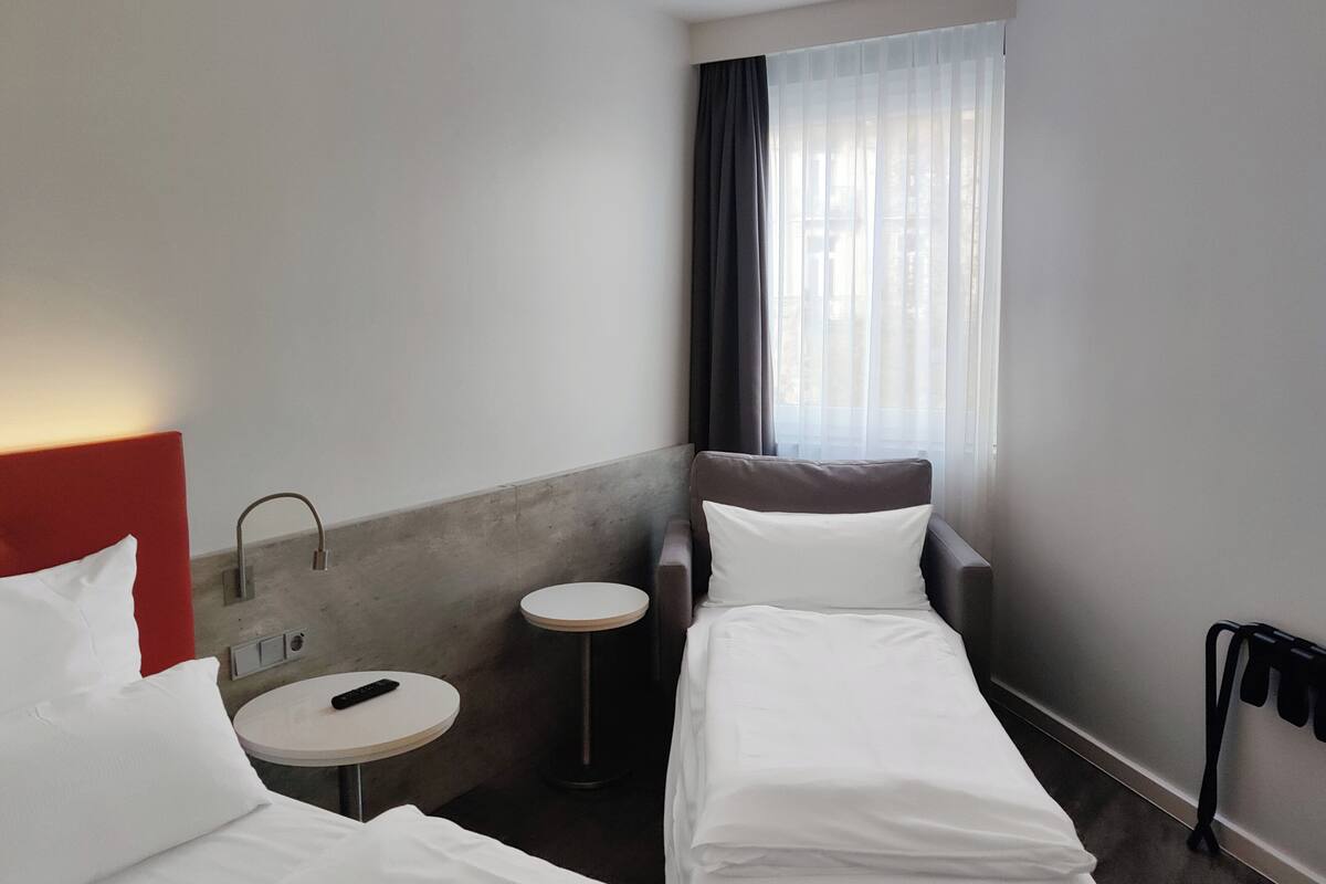 Ein Hotelzimmer mit zwei Einzelbetten, einem runden Tisch, einem roten Kopfteil, einem Fenster mit Vorhängen und einem an der Wand befestigten Telefon.