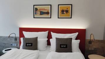 A hotel room with a red headboard, two beds with white bedding, and two pillows with the name "Garnier". There are two framed pictures on the wall, a round table, and a small round stool.