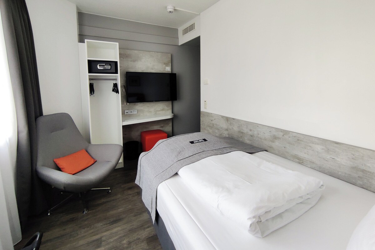 Habitación de hotel con cama, una silla con cojín naranja, armario, televisor de pantalla plana y un taburete rojo.
