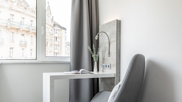 A modern room with a window, a grey chair, a white desk, and a grey curtain. - Garner Hotel Wiesbaden City by IHG (Wiesbaden)