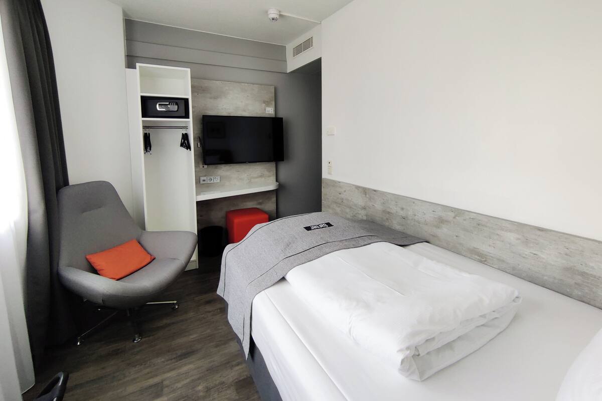 A hotel room with a bed, a chair with an orange cushion, a wardrobe, a flat-screen TV, and a red stool.