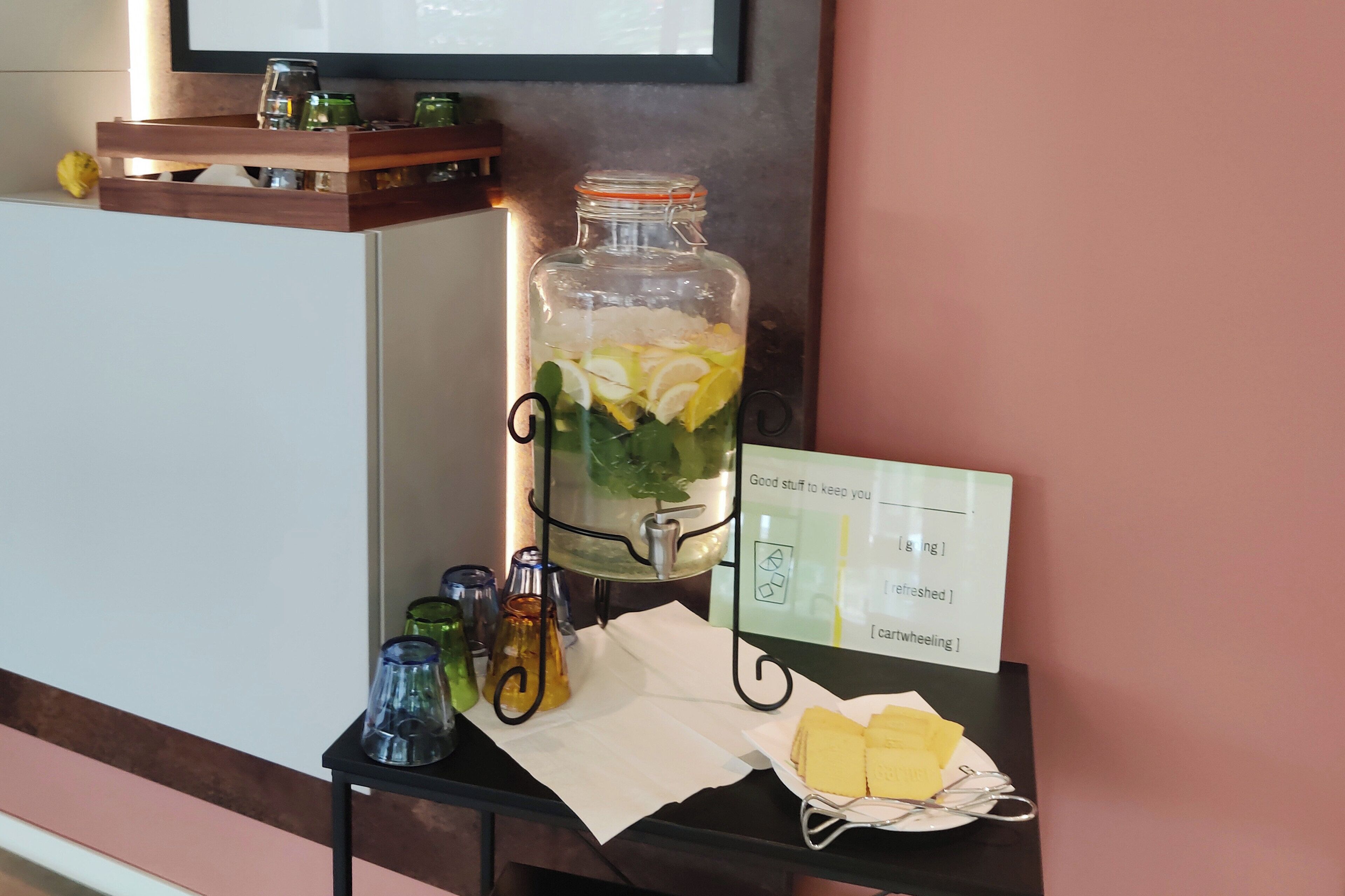 Un dispensador de jugo con un letrero, vasos y una cesta de pan sobre una mesa.
