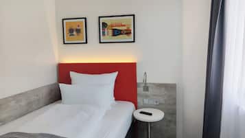 Habitación de hotel con cama, cabecera roja, dos cuadros enmarcados en la pared, mesita de noche con control remoto y baño moderno con cortina de ducha.