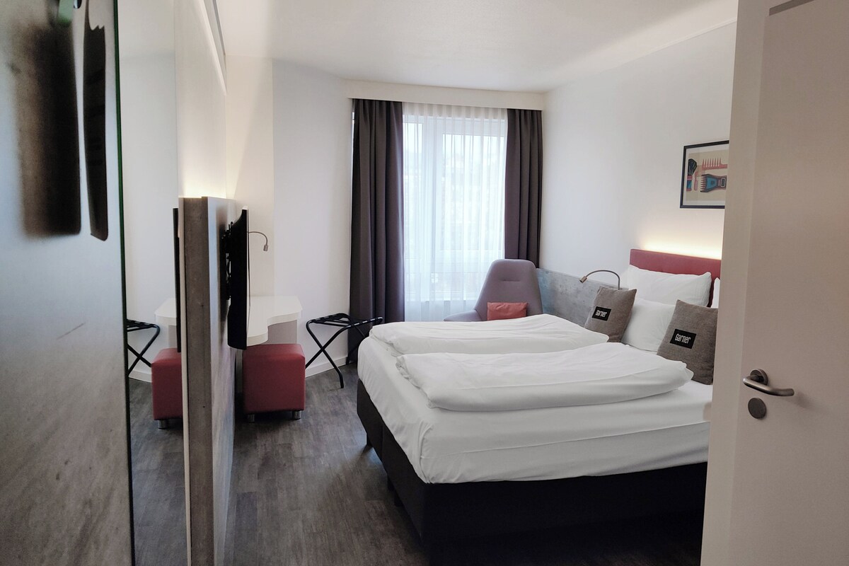 Una habitación de hotel con cama, escritorio, silla y ventana con cortinas.
