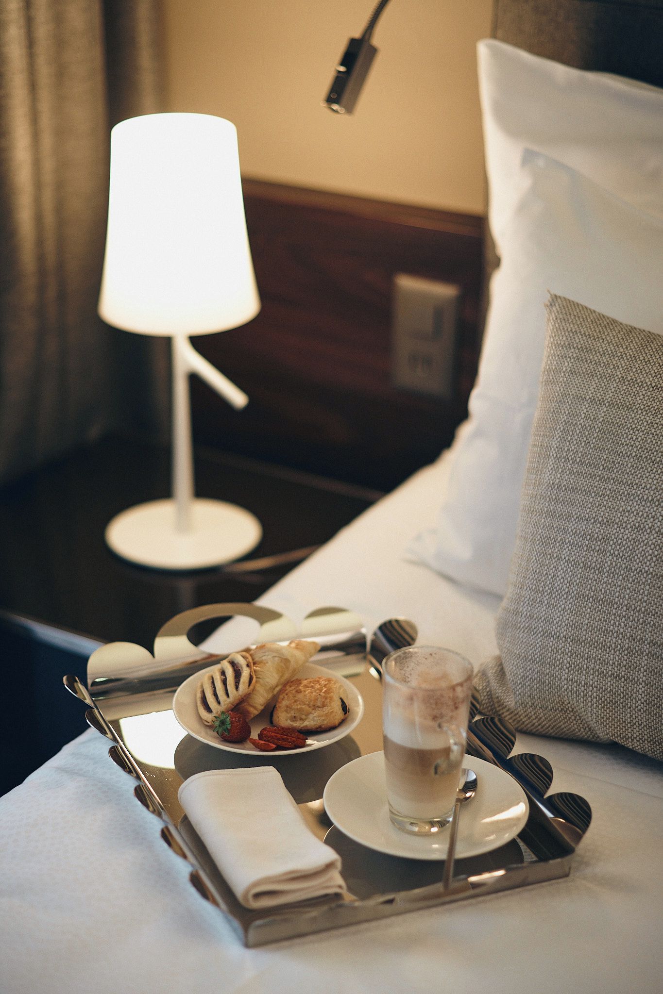 En pænt anrettet bakke med bagværk, et glas drikke og bestik på en hotelseng.