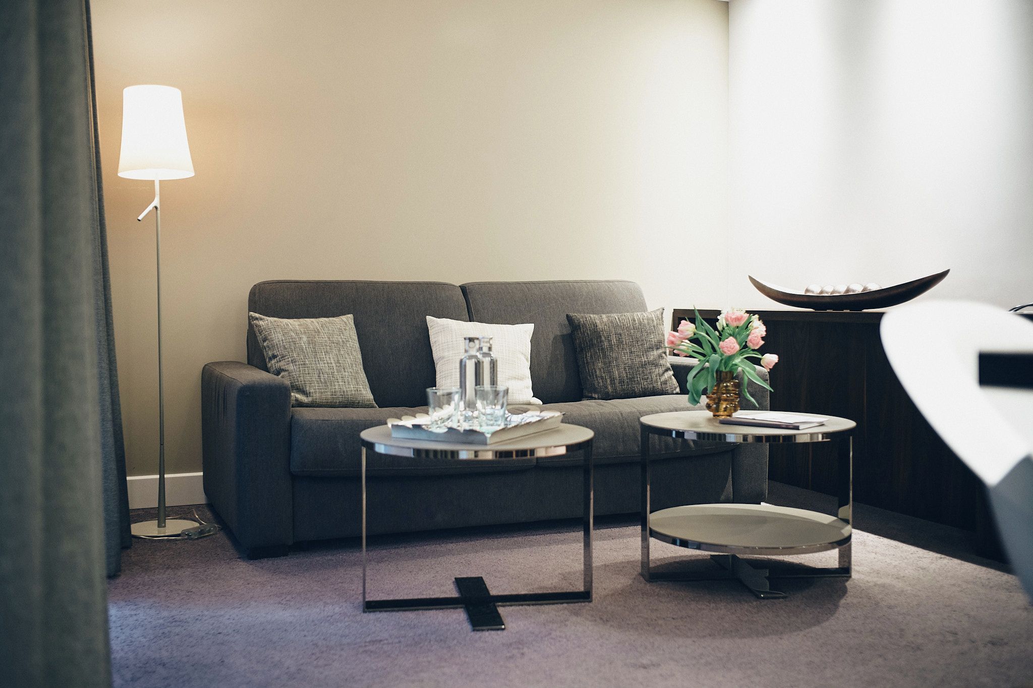 Ein modernes Wohnzimmer mit einem grauen Sofa, zwei Beistelltischen, einer Stehlampe und einer Vase mit Blumen.