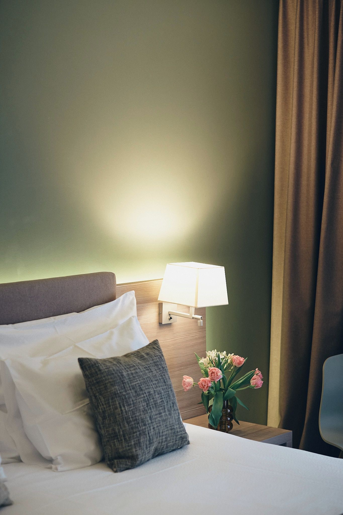 Een hotelkamer met een bed, kussens, een nachtkastje met een lamp en een vaas met bloemen.