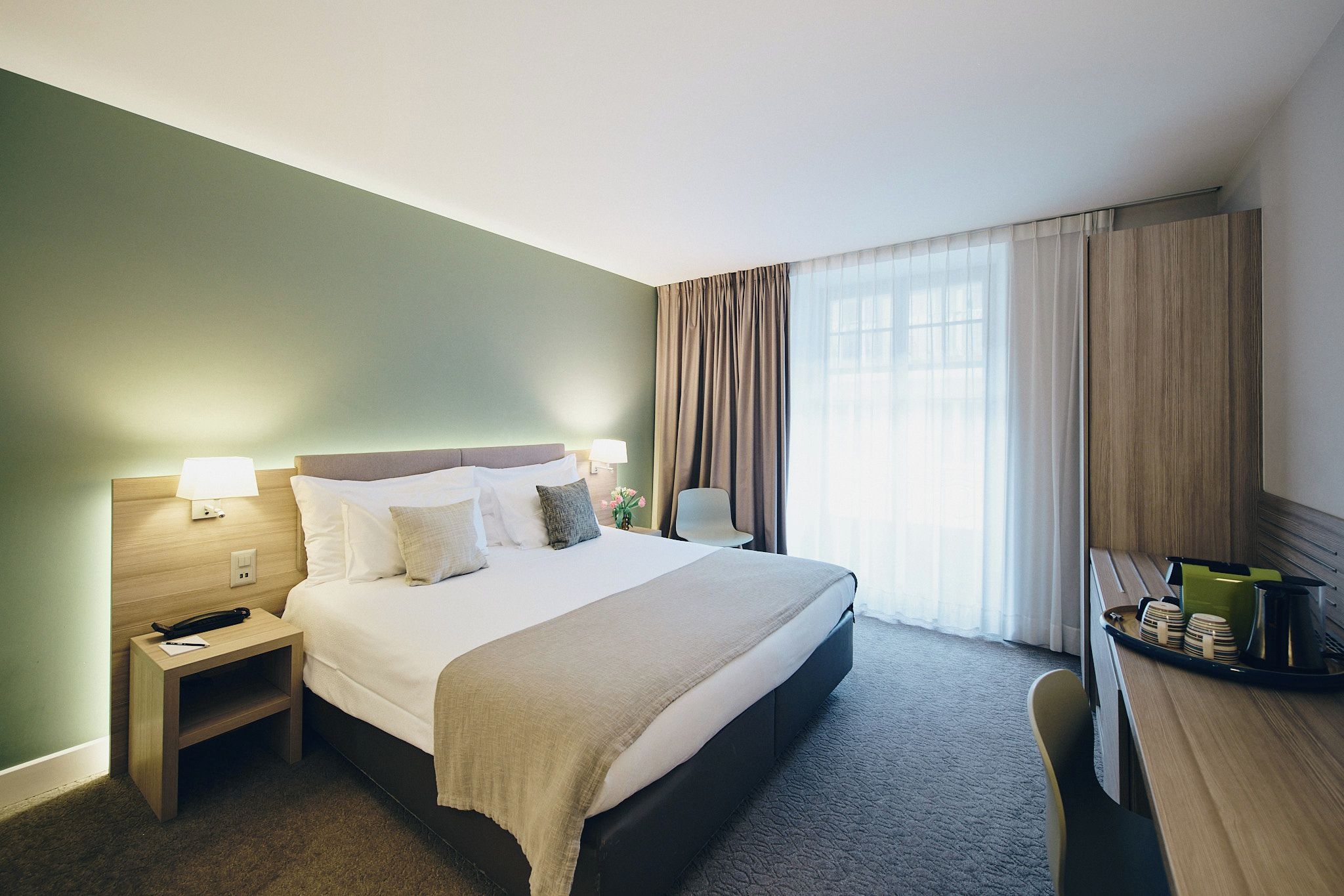 Ein Hotelzimmer mit einem Bett, Nachttischen, einem Schreibtisch und einem fensterbepflanzten Fenster.