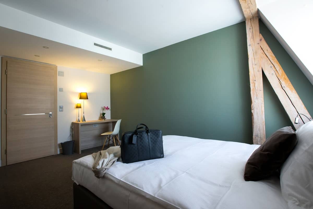 Ett hotellrum med en säng, ett skrivbord, en stol, en lampa och en väska.