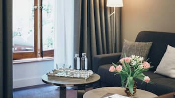 Une chambre d’hôtel avec un canapé, deux tables basses, une lampe, une fenêtre avec des rideaux et un vase de fleurs.