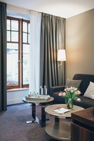 A hotel room with a sofa, two coffee tables, a lamp, a window with curtains, and a vase with flowers.