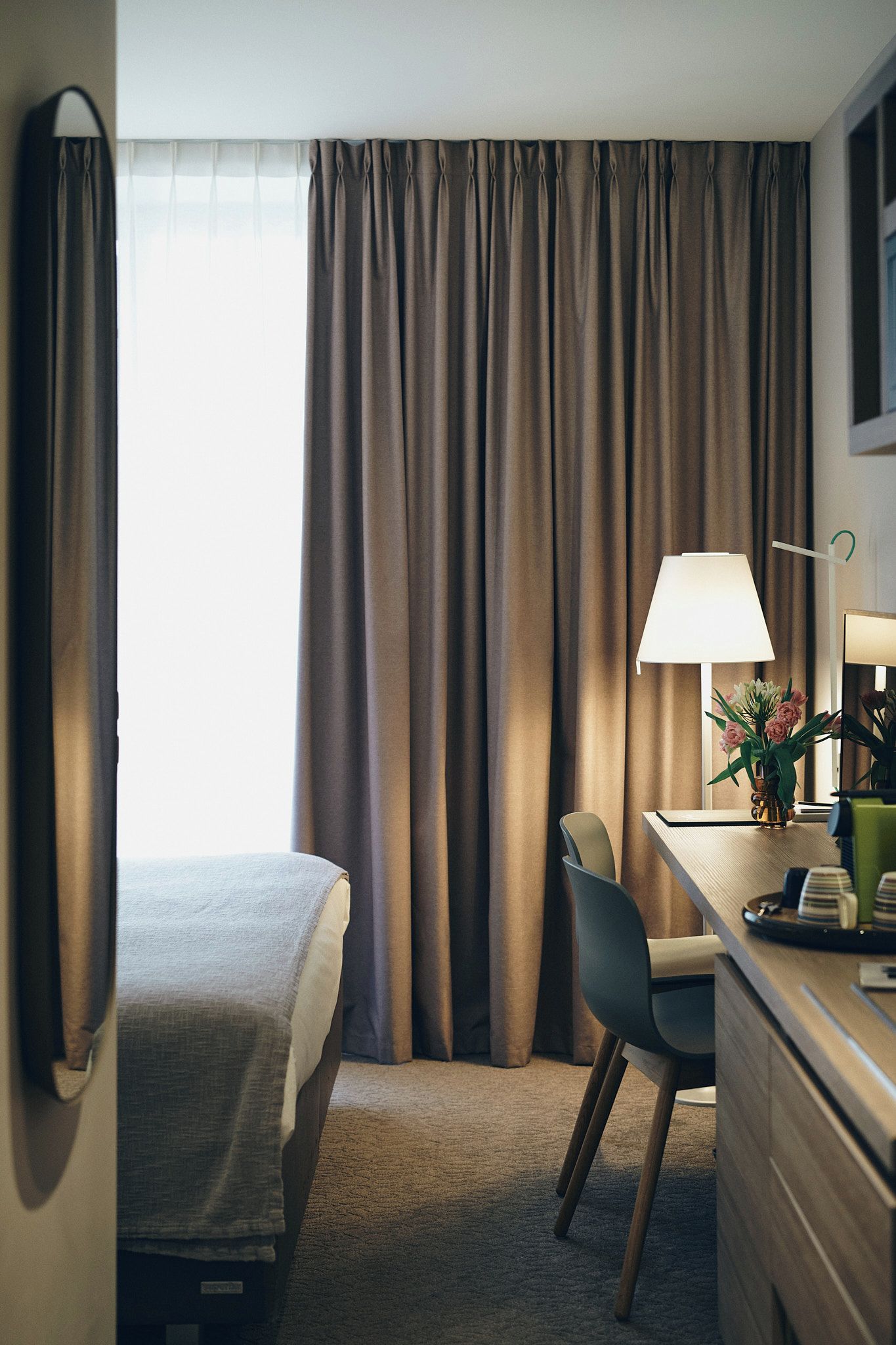Een hotelkamer met een bed, een bureau met een lamp, een stoel en uitzicht door gordijnen.