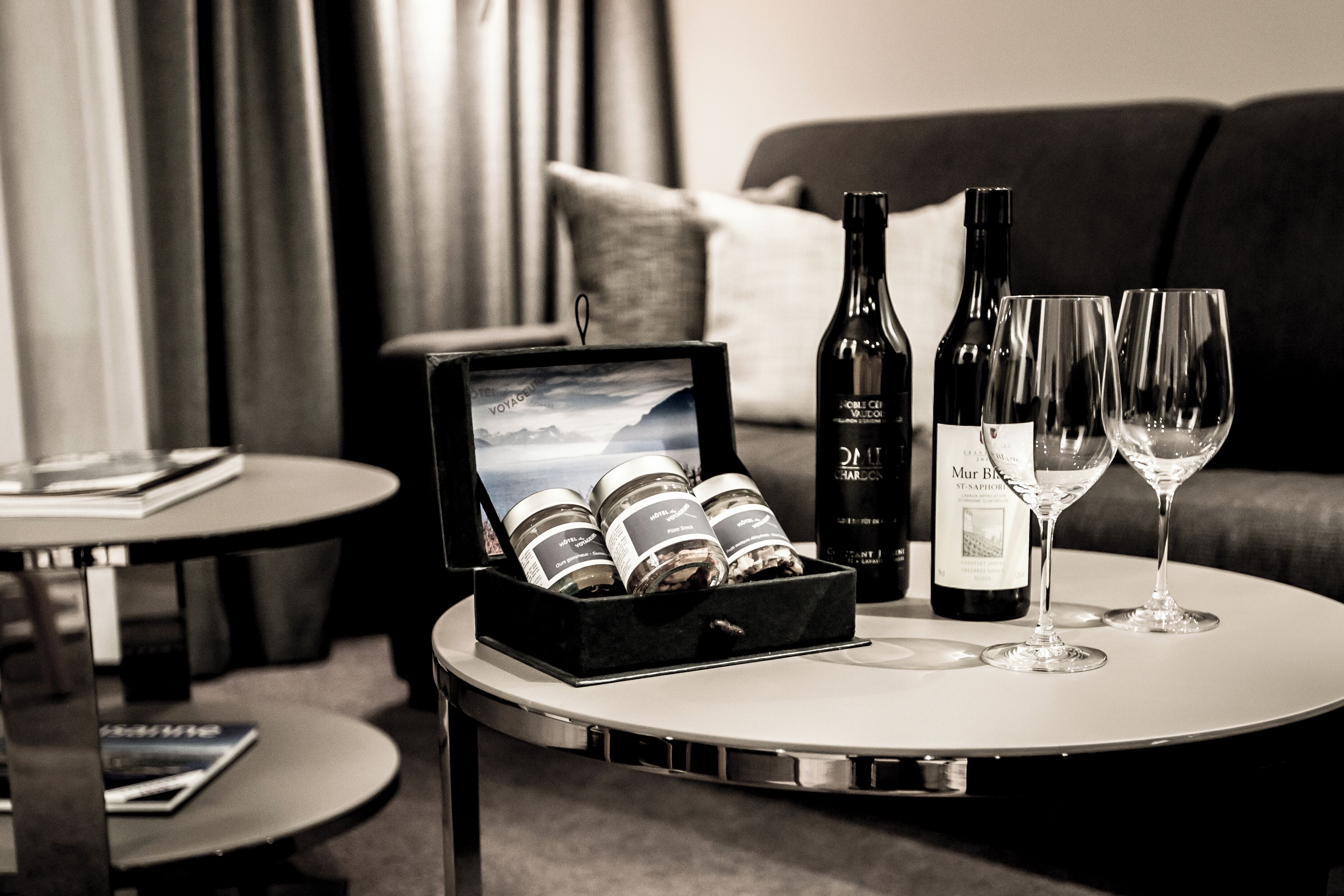Habitación de hotel con una mesa redonda que presenta una botella de vino, dos copas de vino y una caja de bombones.