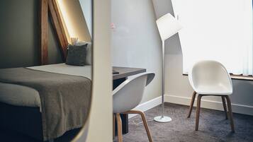 A modern hotel room with a bed, a desk, a chair, and a floor lamp.