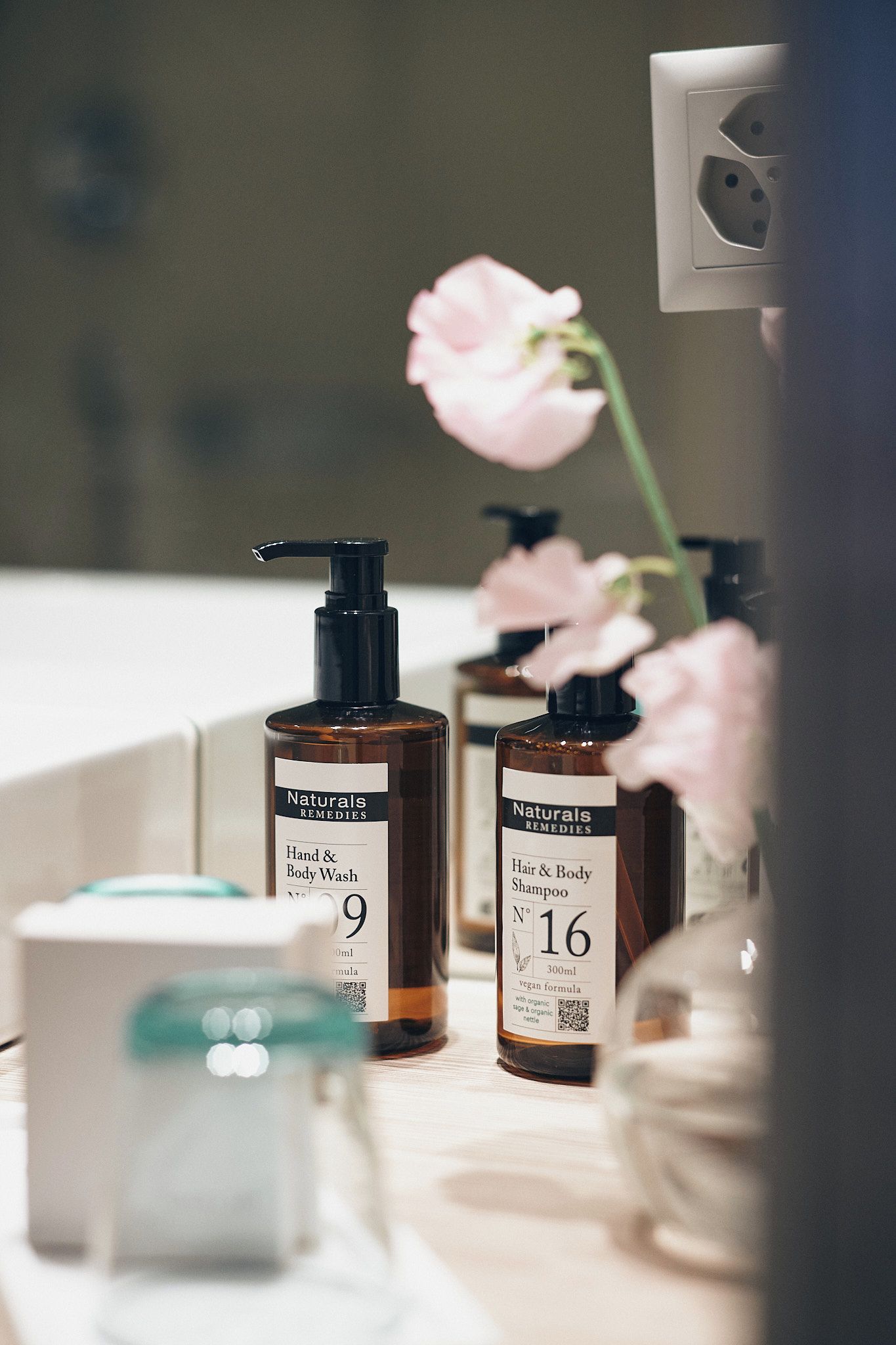 A bathroom vanity with two bottles of hand and body wash, a hair and body shampoo, and a vase with a pink flower.