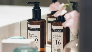 A bathroom vanity with two bottles of hand and body wash, a hair and body shampoo, and a vase with a pink flower.