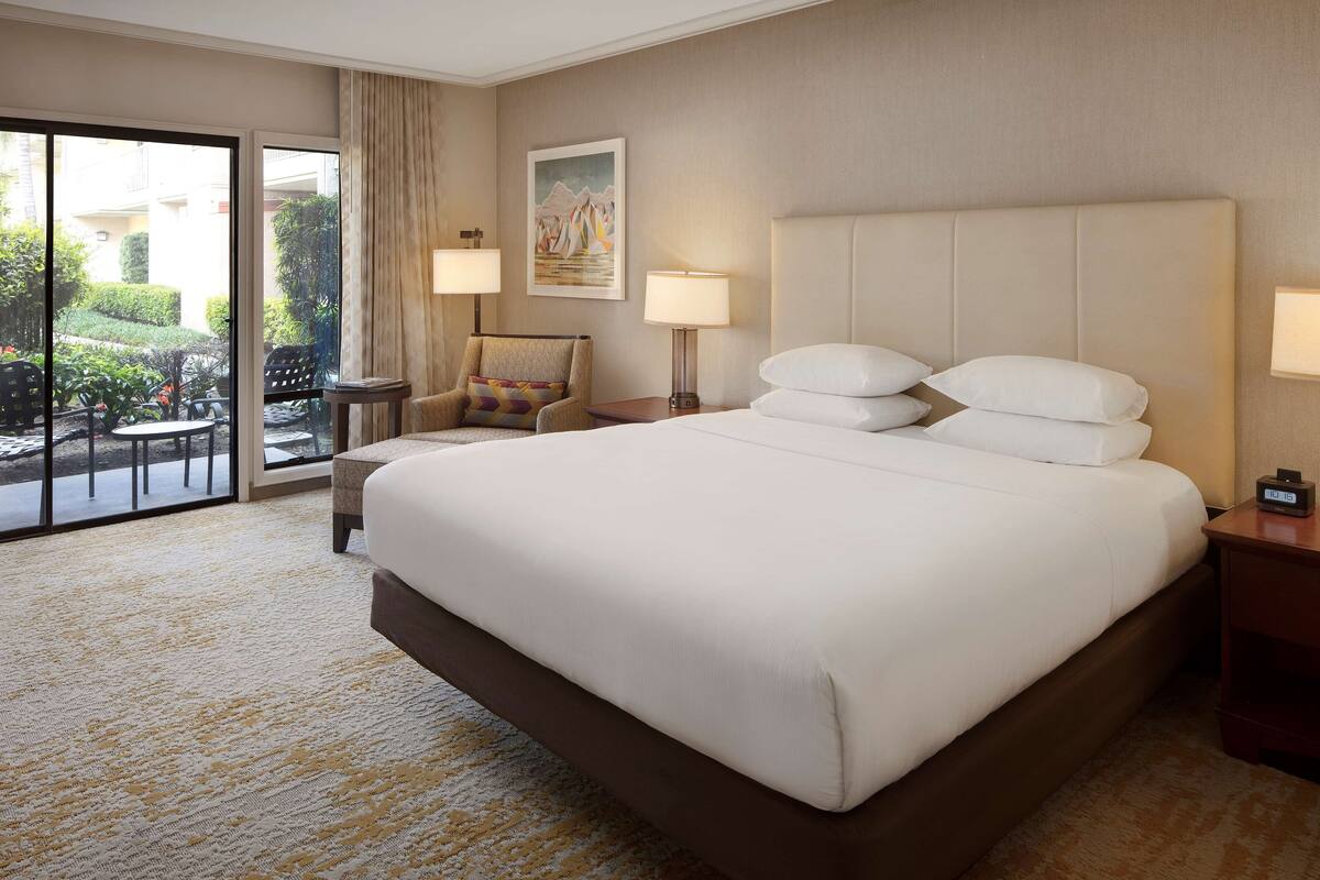 Ein Hotelzimmer mit einem großen Bett, zwei Nachttischlampen, einem Sessel, einem kleinen Tisch und Blick ins Freie durch eine Schiebetür.