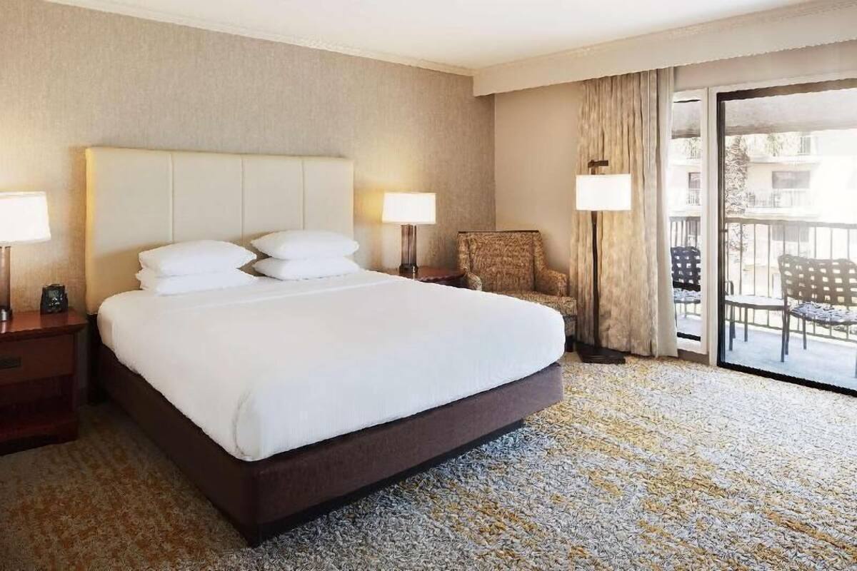 Habitación de hotel con una cama grande, dos lámparas de noche, una silla y un balcón con mobiliario exterior.