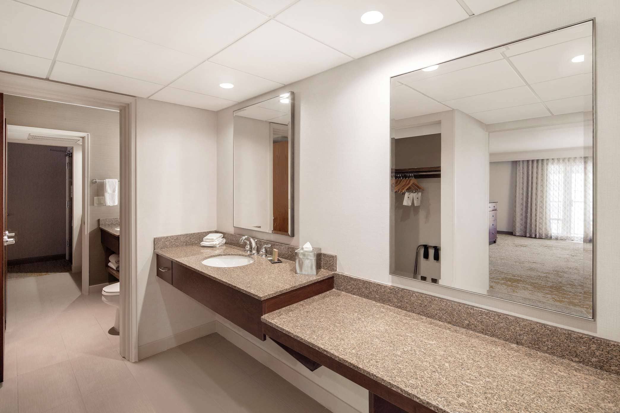 Un baño moderno con lavamanos doble, un espejo grande y un vestidor visible a través de una puerta abierta.