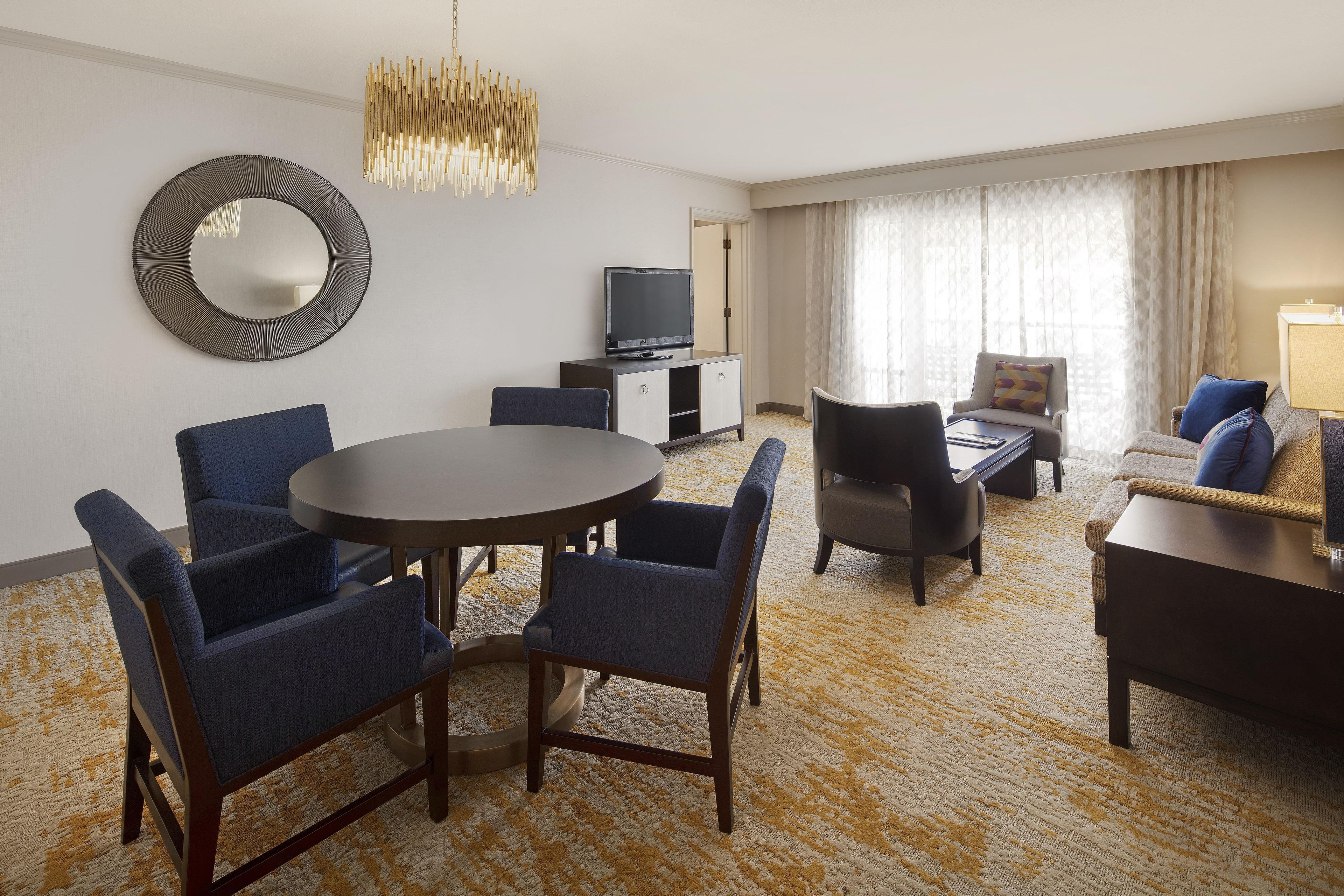 Habitación de hotel con una mesa de comedor redonda, dos sillas azules, un sofá, un televisor y un espejo grande en la pared.