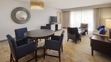 Habitación de hotel con una mesa de comedor redonda, dos sillas azules, un sofá, un televisor y un espejo grande en la pared.