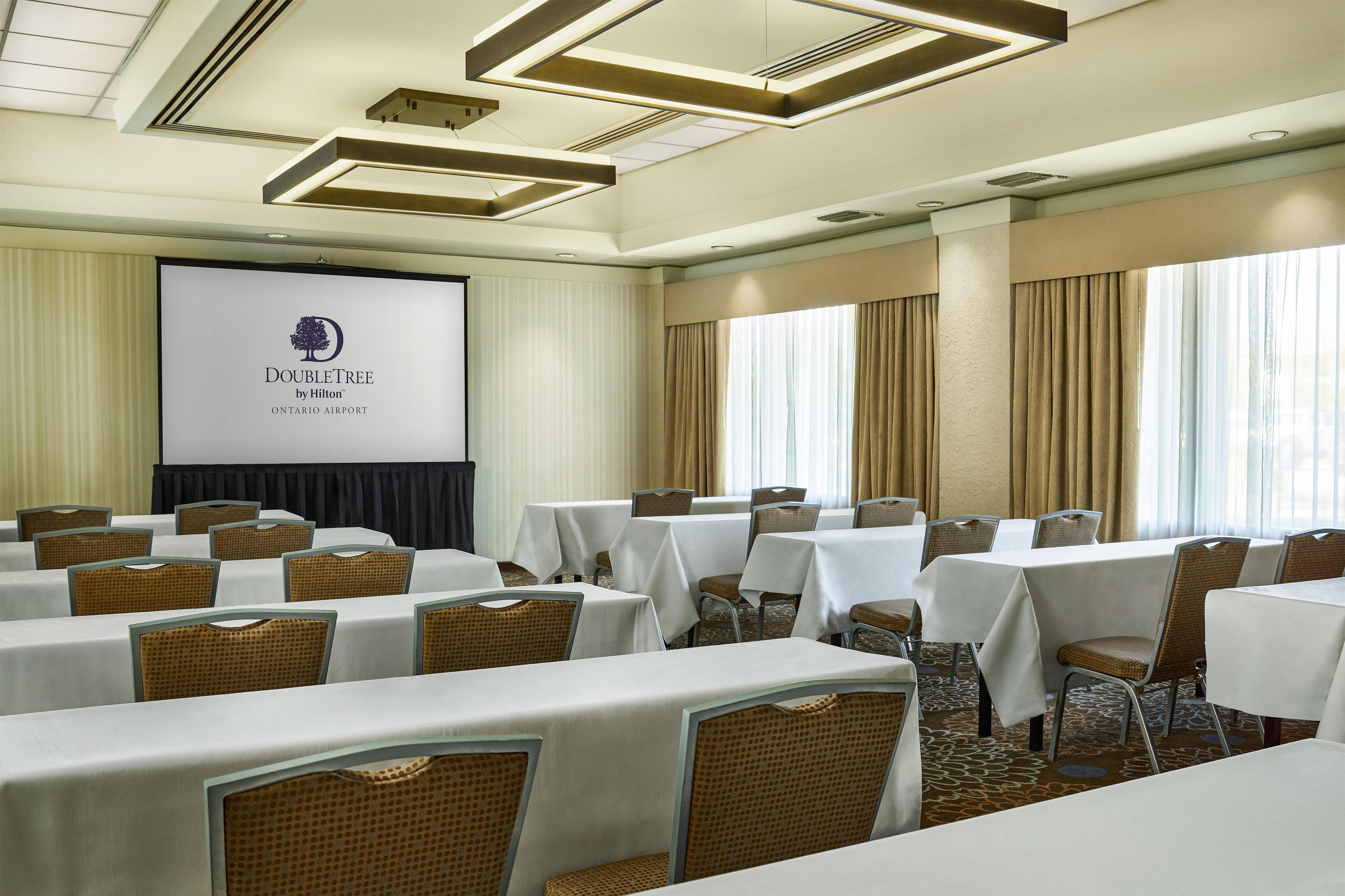 A conference room with tables and chairs, a projection screen, and a logo for DoubleTree by Hilton at Ontario Airport.