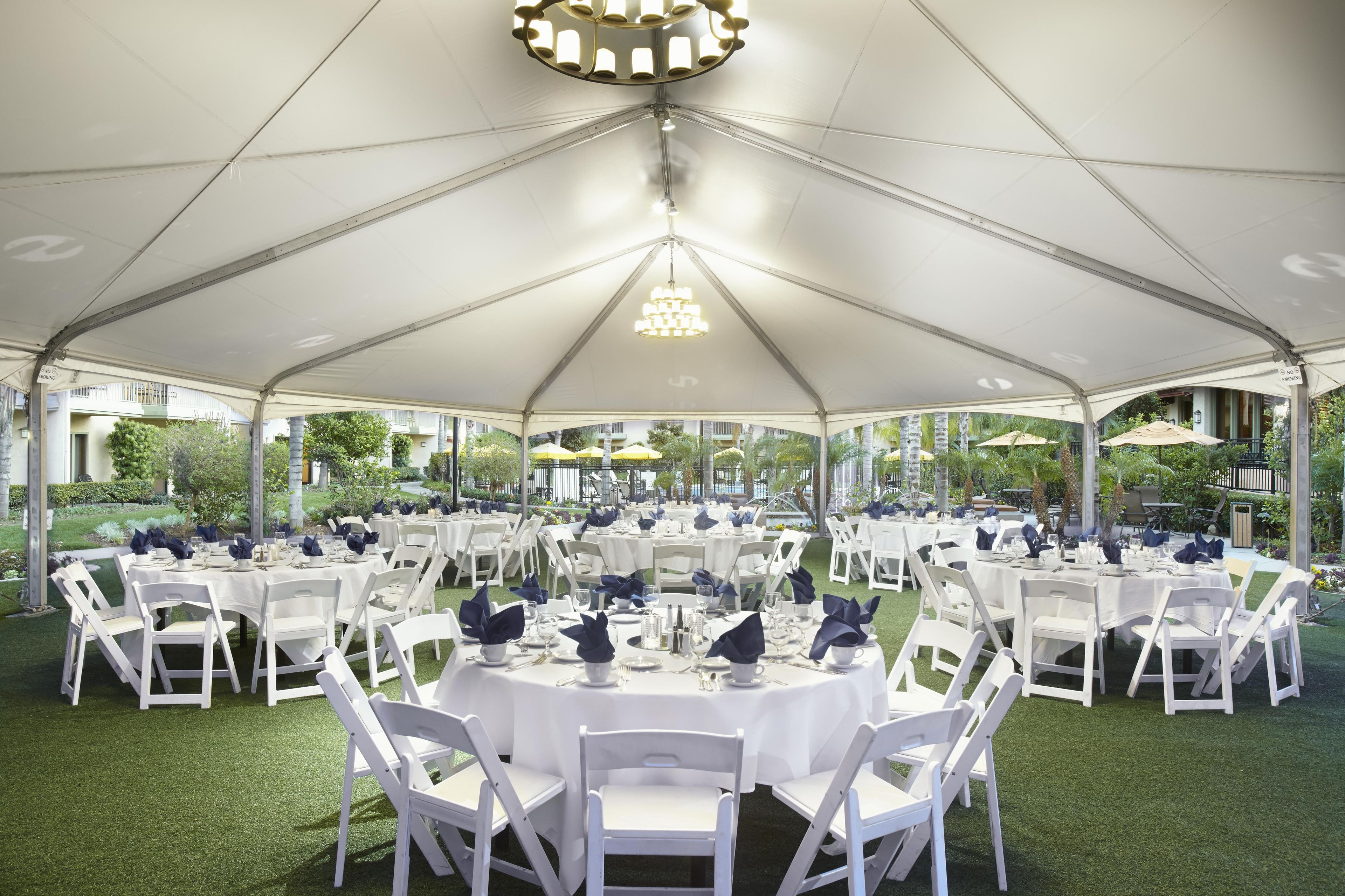 A tented outdoor dining area with round tables and white chairs, surrounded by greenery.
