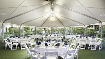 A tented outdoor dining area with round tables and white chairs, surrounded by greenery.