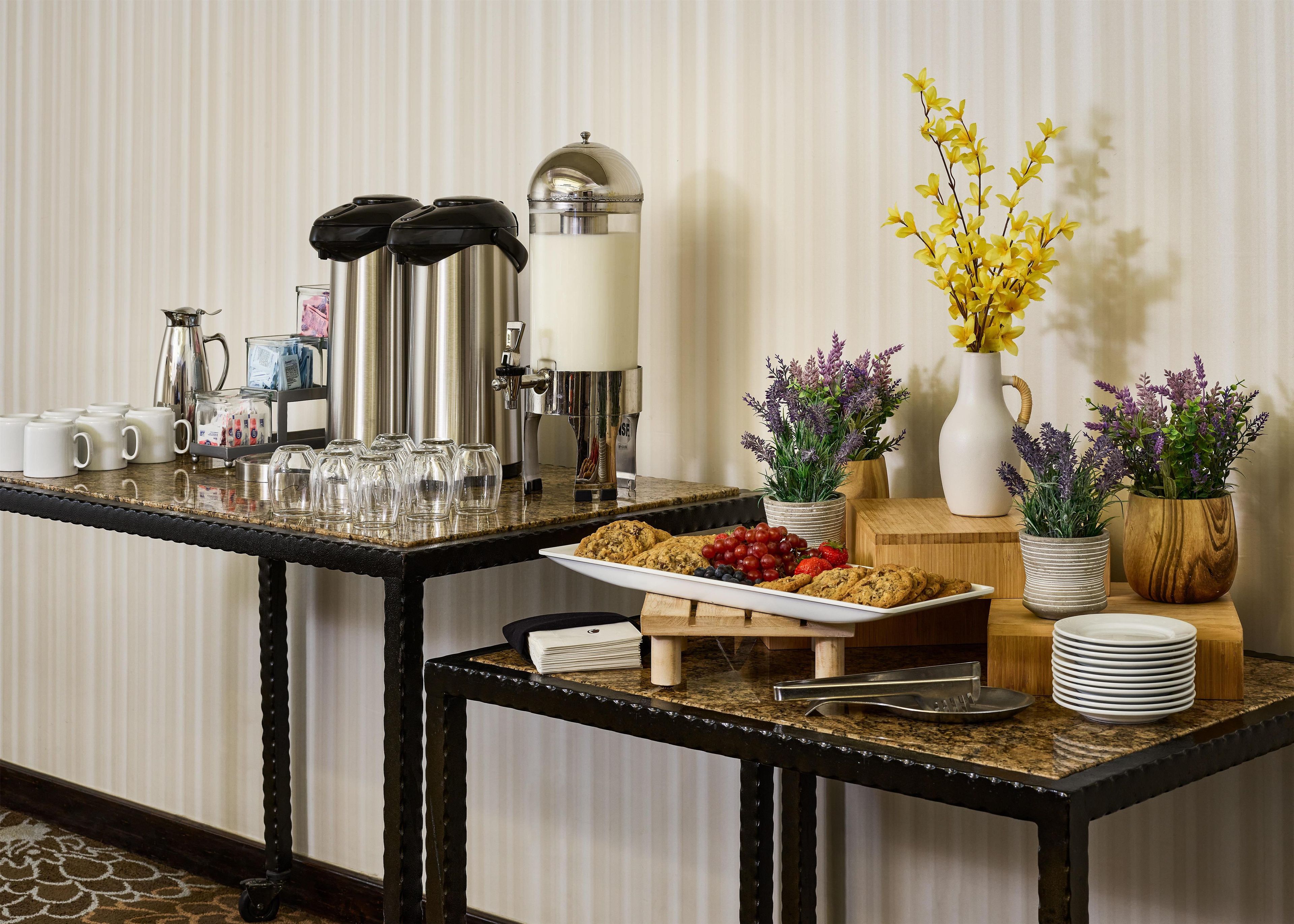 A breakfast buffet setup with coffee machines, a fruit platter, cookies, and decorative plants.