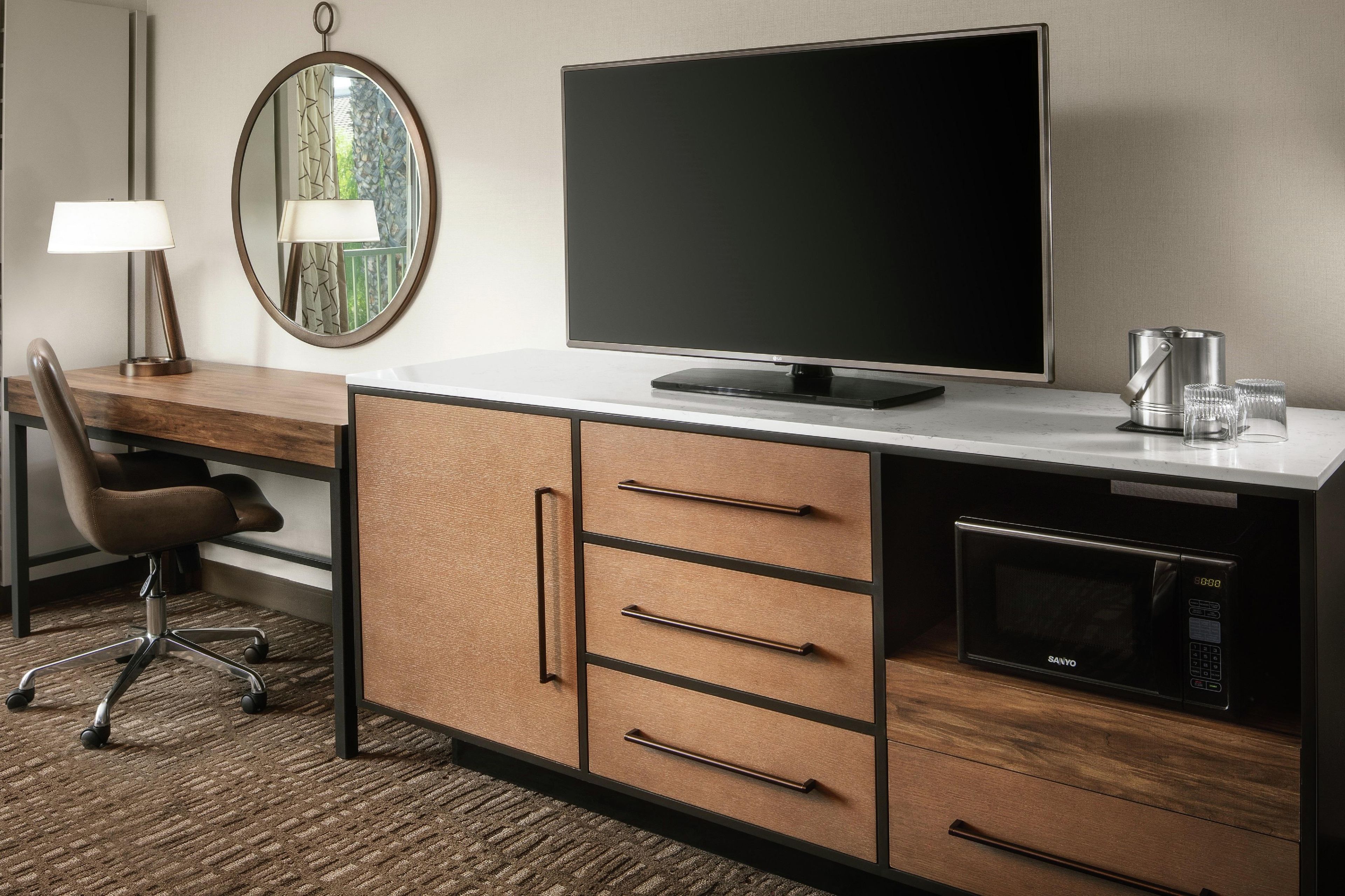 A hotel room with a flat-screen TV, a microwave, a coffee maker, a desk with a chair, and a mirror.