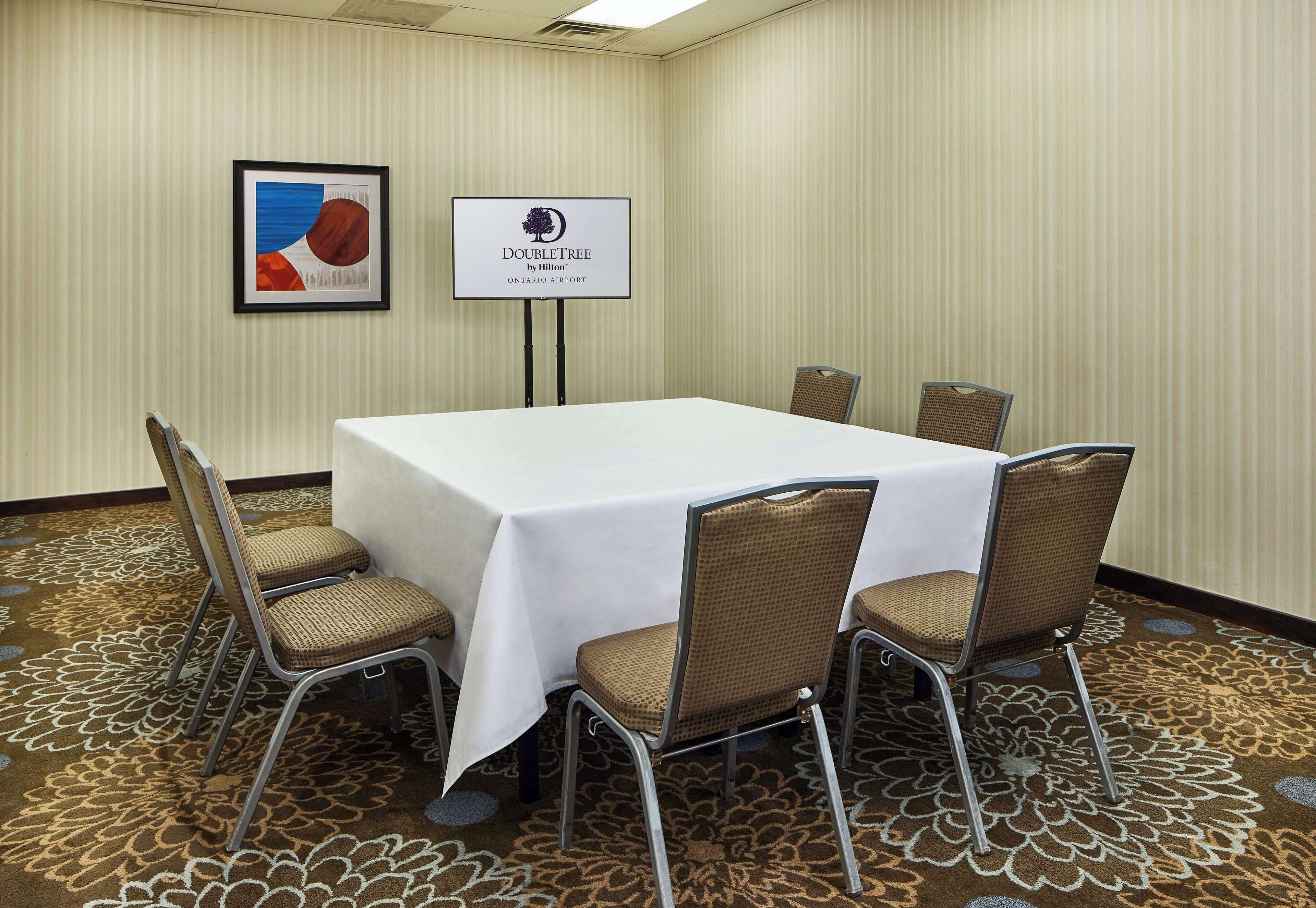 A conference room with a white table, six wicker chairs, a framed abstract painting, and a sign for DoubleTree by Hilton at Ontario Airport.