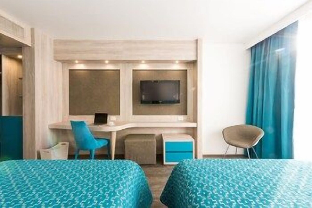 Ein Hotelzimmer mit zwei Betten, einem Schreibtisch mit Stuhl, einem Fernseher und einem Fenster mit Vorhängen.