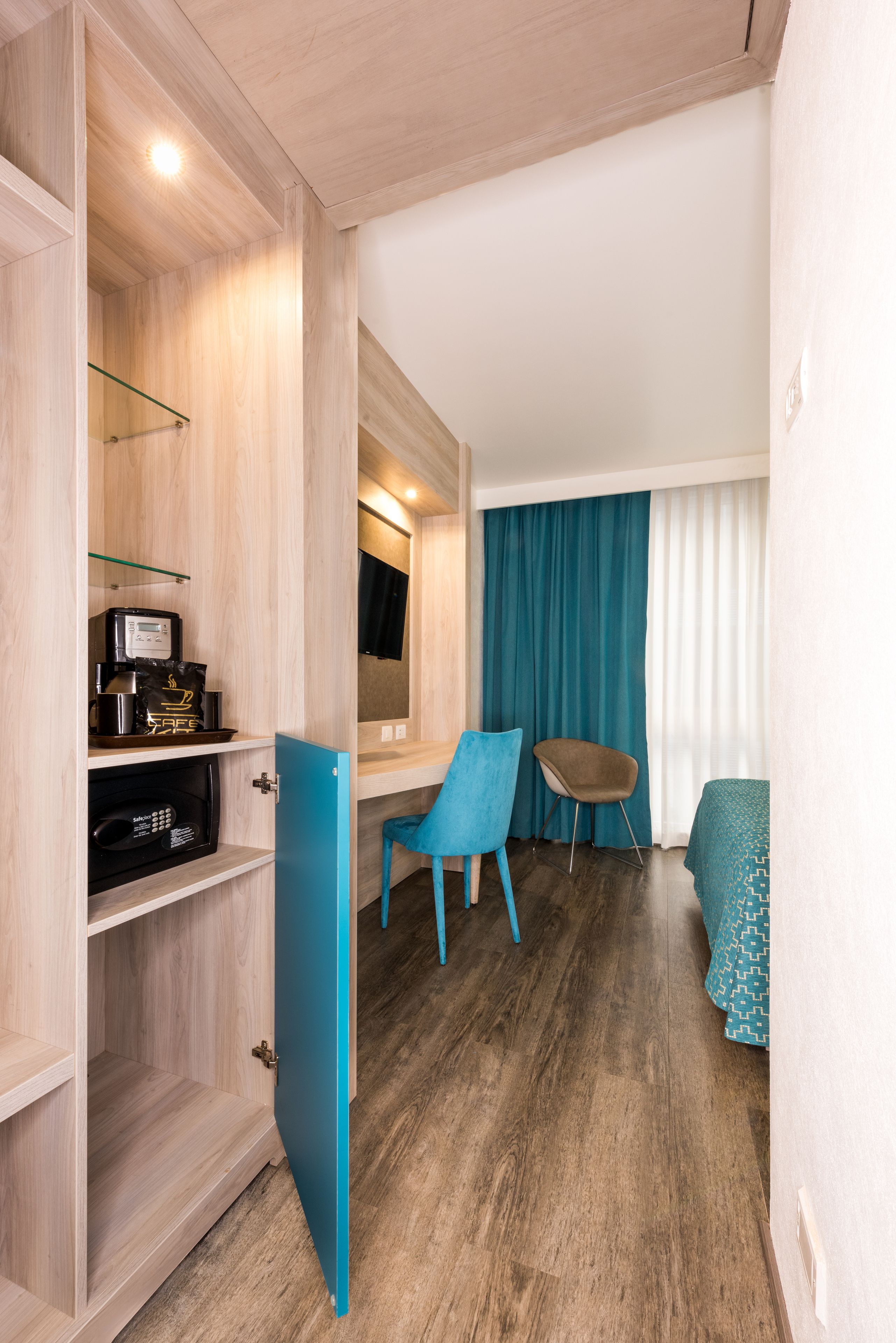 A compact room with a wooden wardrobe, a small table with a coffee machine, a blue chair, and a bed with a patterned bedspread.