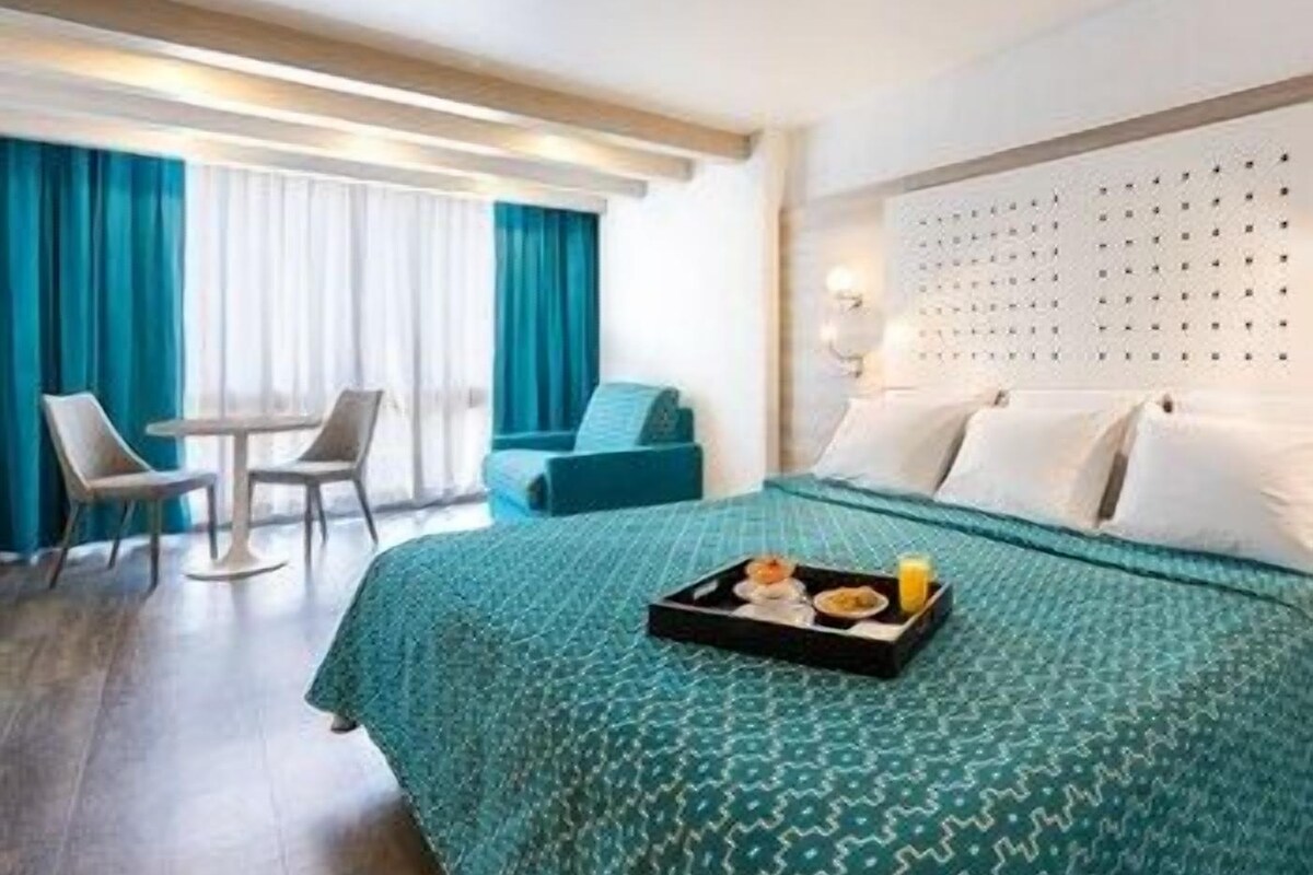 Ein Hotelzimmer mit Bett, Esstisch, Stühlen, einem Fenster mit Vorhängen und einem Tablett mit Frühstücksangeboten.
