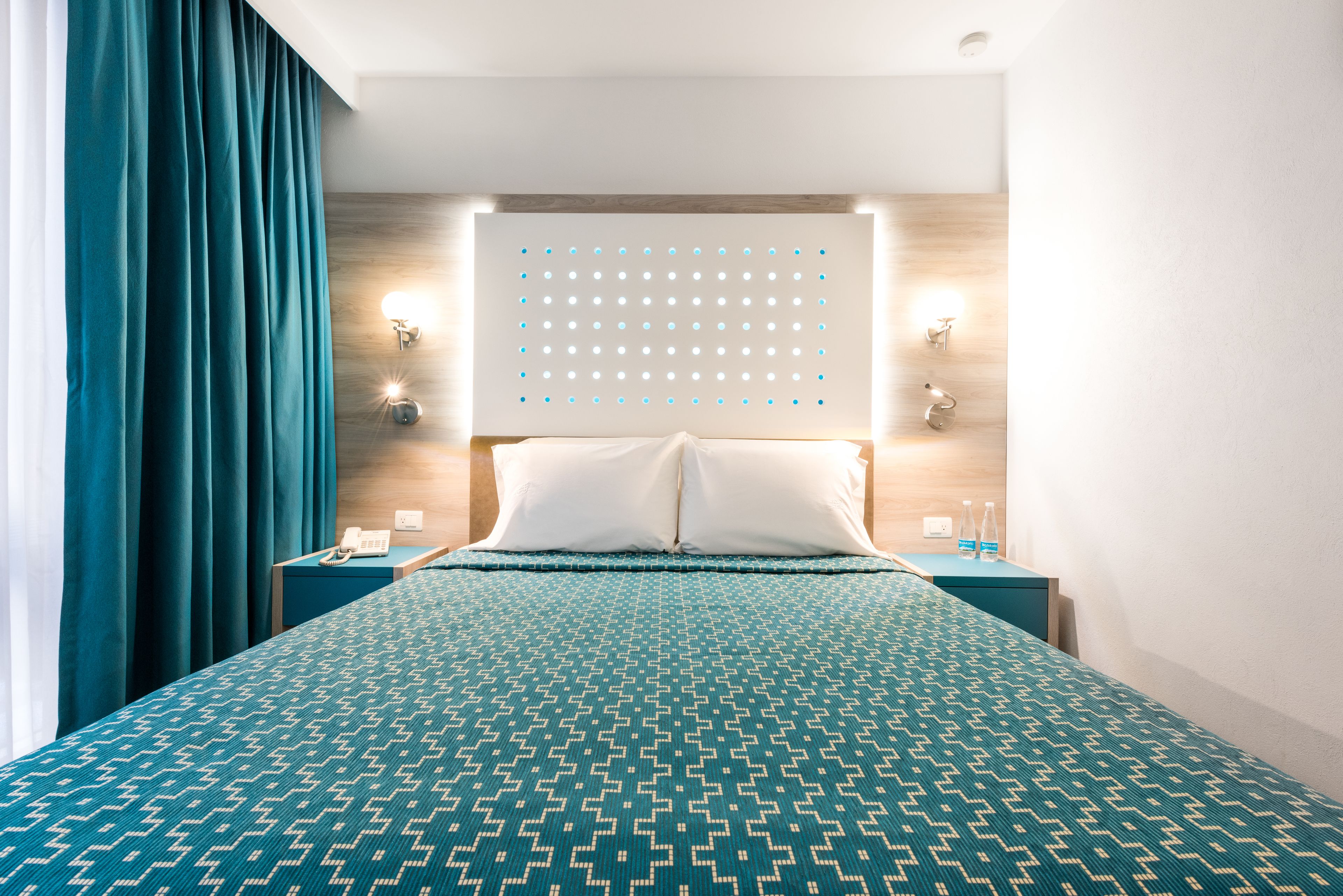 A hotel room with a large bed, teal curtains, a wooden headboard, two bedside tables with lamps, and a wall-mounted light fixture.