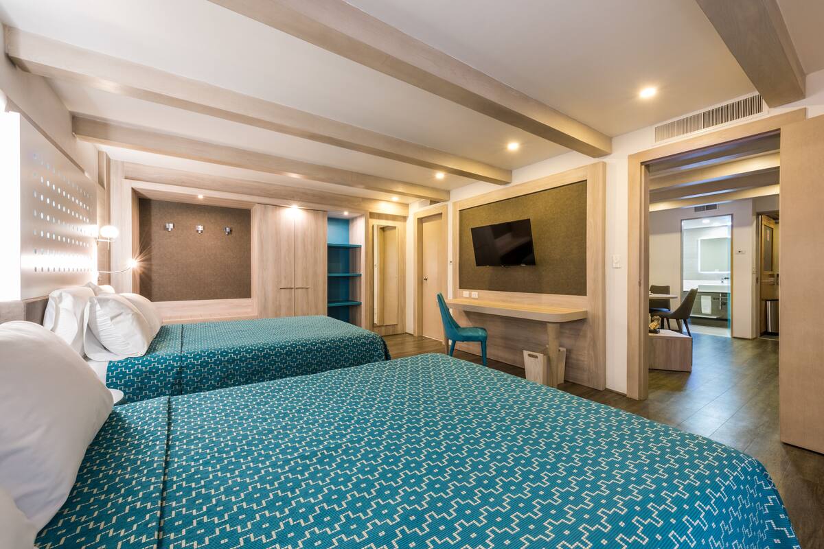 Habitación de hotel con dos camas, televisión, escritorio y baño visible a través de una puerta abierta.
