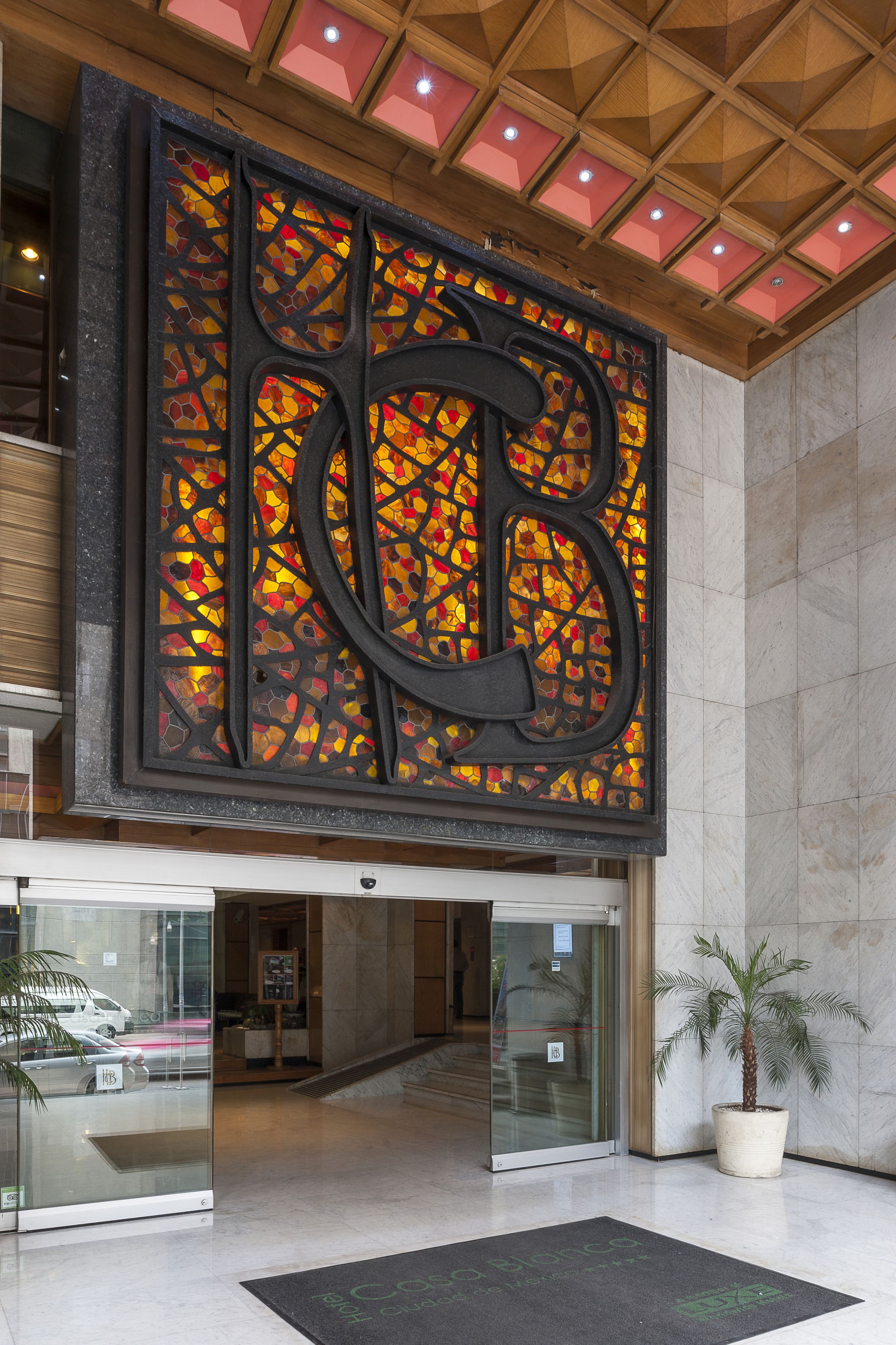 Una entrada de edificio grande y moderna, con un destacada fachada de vidrio, una gran letra decorativa 'B' en la pared y una palmera en maceta.