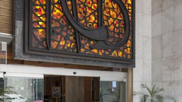 Una entrada de edificio grande y moderna, con un destacada fachada de vidrio, una gran letra decorativa 'B' en la pared y una palmera en maceta.