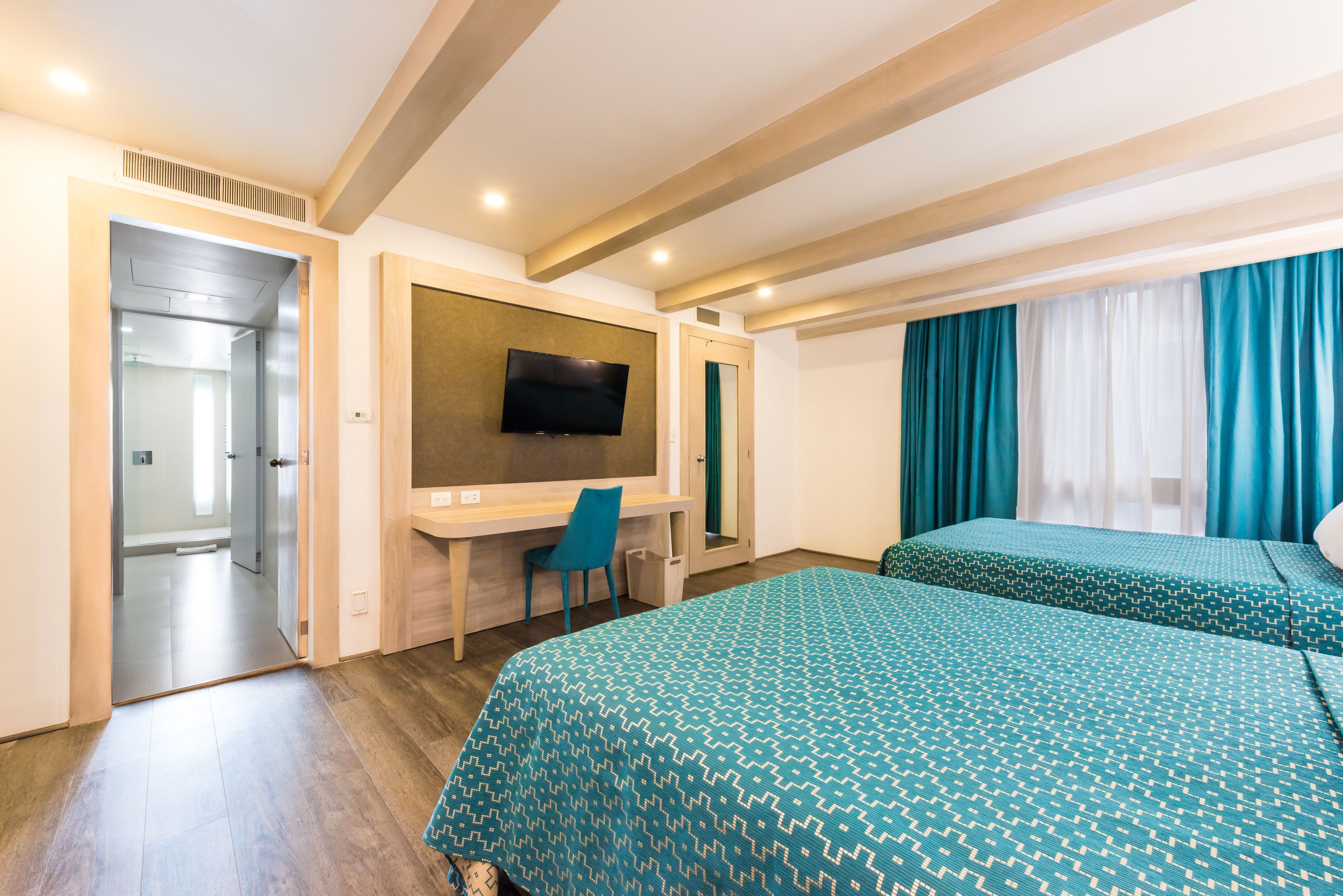 Habitación de hotel con techo de madera, un escritorio con silla, cama con ropa de cama estampada y televisor montado en la pared.