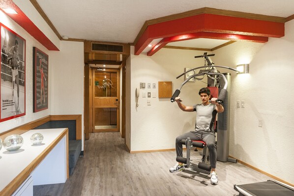 Un gimnasio con una persona usando una máquina de poleas sentada, suelo de madera y un mostrador de recepción con adornos.
