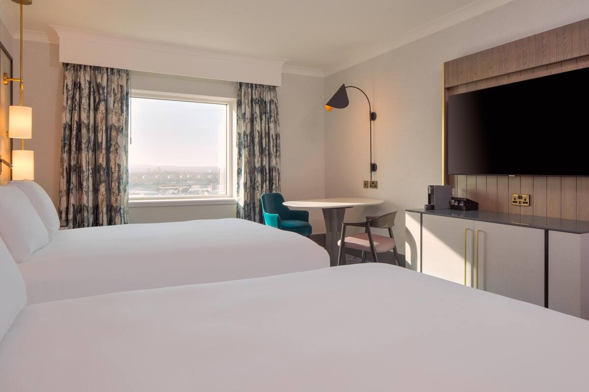 A hotel room with two beds, a desk, a chair, a TV, and a window with curtains.
