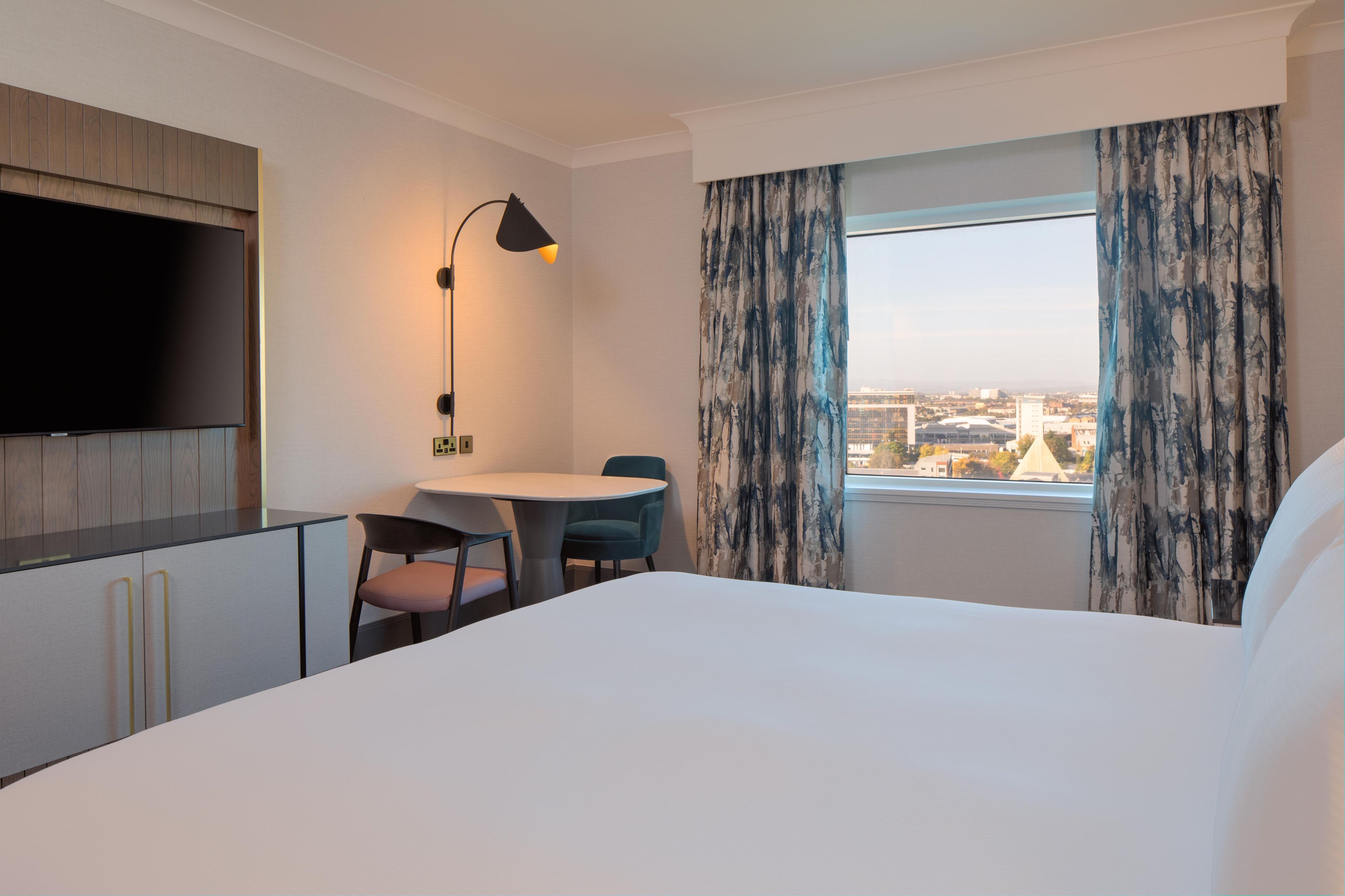 Habitación de hotel con cama, escritorio, silla, televisor, lámpara y ventana con cortinas.