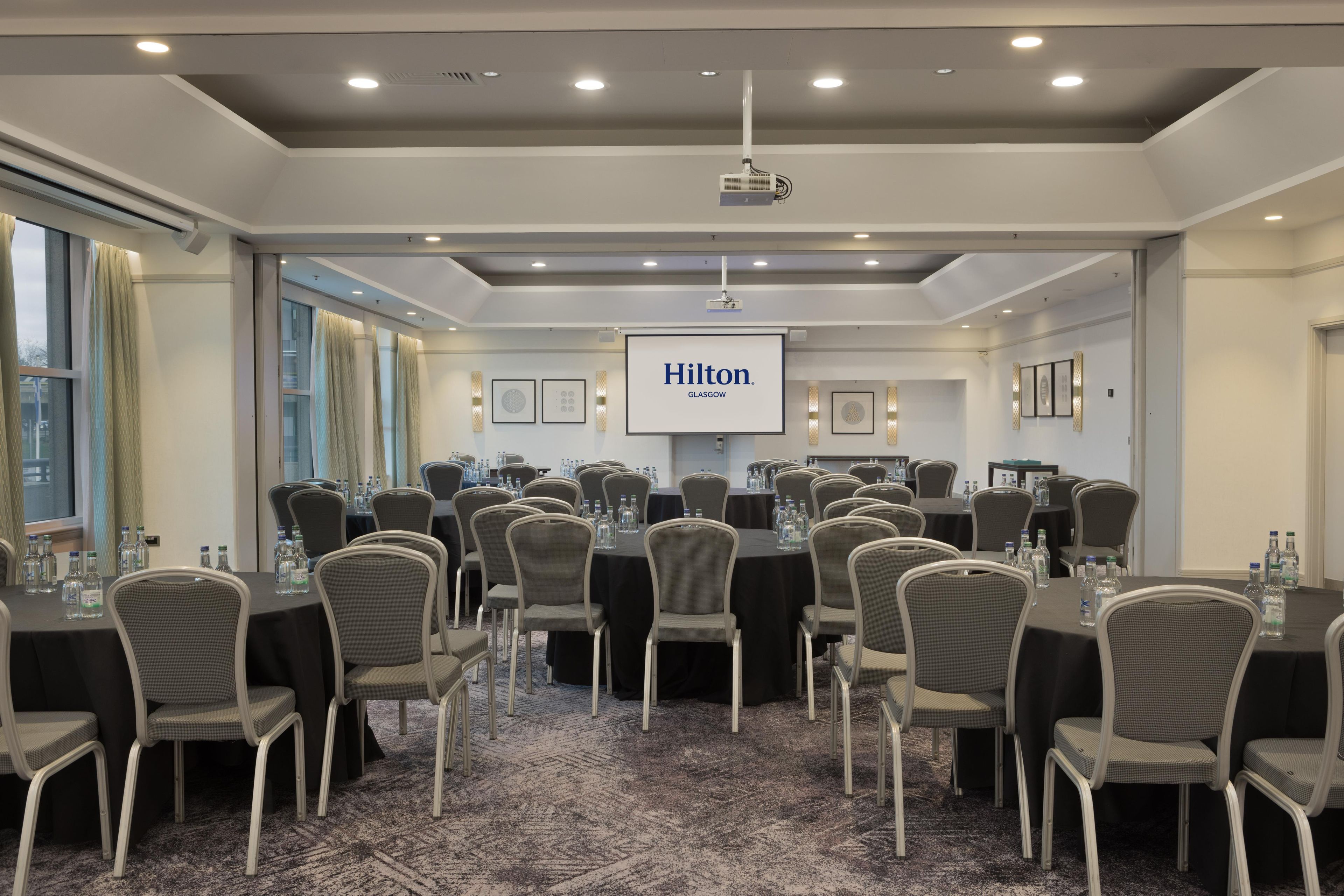 Una sala de conferencias con varias mesas y sillas, una pantalla de proyector mostrando 'Hilton Glasgow' y un gran ventanal con cortinas.