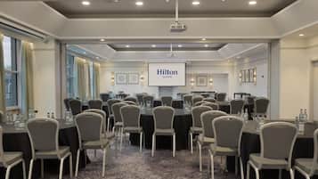 Una sala de conferencias con varias mesas y sillas, una pantalla de proyector mostrando 'Hilton Glasgow' y un gran ventanal con cortinas.